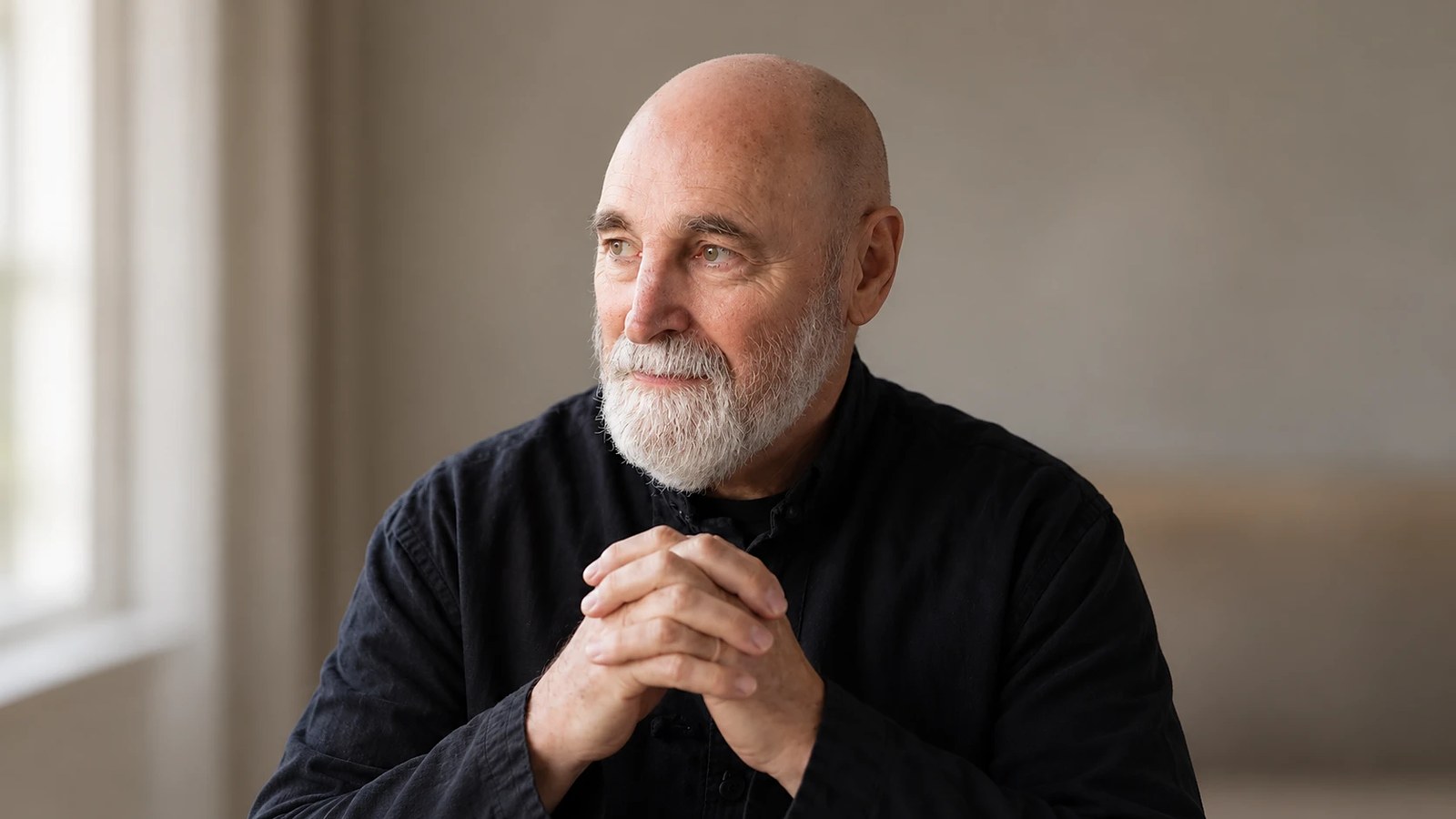 Rev. Bill McDonald seated by a window in soft natural light, hands clasped, gazing thoughtfully into the distance.
